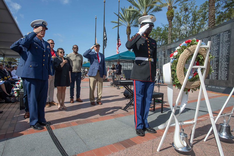 Several people and Marine saluting a wreath laid at the Memorial Wall during the Memorial Day 2026 service